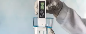 pH-Meter in den Händen mit Handschuhen, Glas Wasser auf blauem Hintergrund. Messung der Eigenschaften von Trinkwasser.
