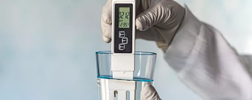 pH-Meter in den Händen mit Handschuhen, Glas Wasser auf blauem Hintergrund. Messung der Eigenschaften von Trinkwasser.